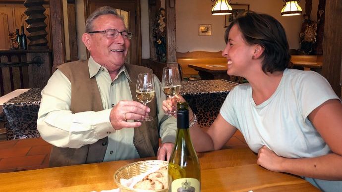 Ein älterer Herr und eine junge Frau stoßen mit Weingläsern an.