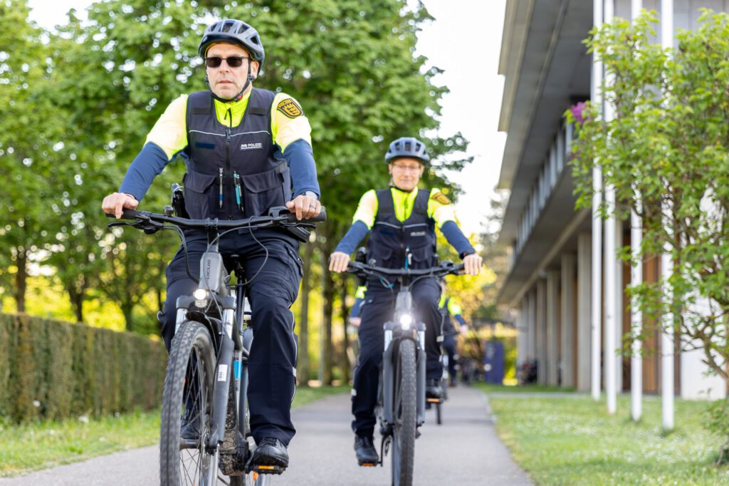 Ein Fahrradpolizist mit Sonnenbrille auf dem Fahrrad draußen, im Hintergrund eine Fahrradpolizistin.
