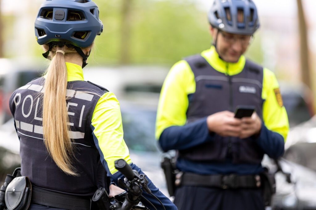 Fahrradpolizistin steht mit Rücken zu Kamera, Fahrradpolizist im Hintergrund tippt etwas in sein Handy.